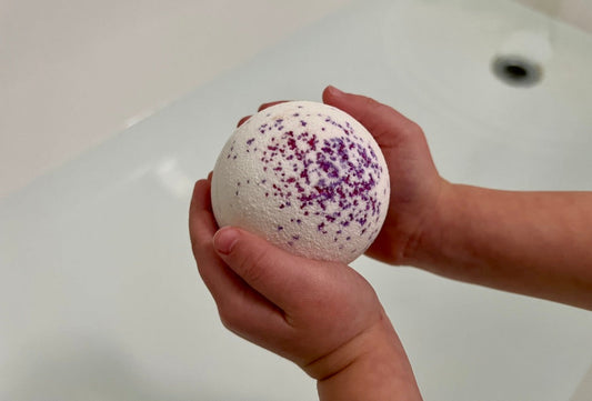 Child holding a dye-free bath bomb for kids with purple sprinkles over bathtub